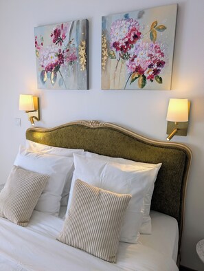 Un lit bien fait, avec du linge de lit blanc et des oreillers beige, flanqué de deux lampes fixées au mur, dotées d’abat-jour blancs.