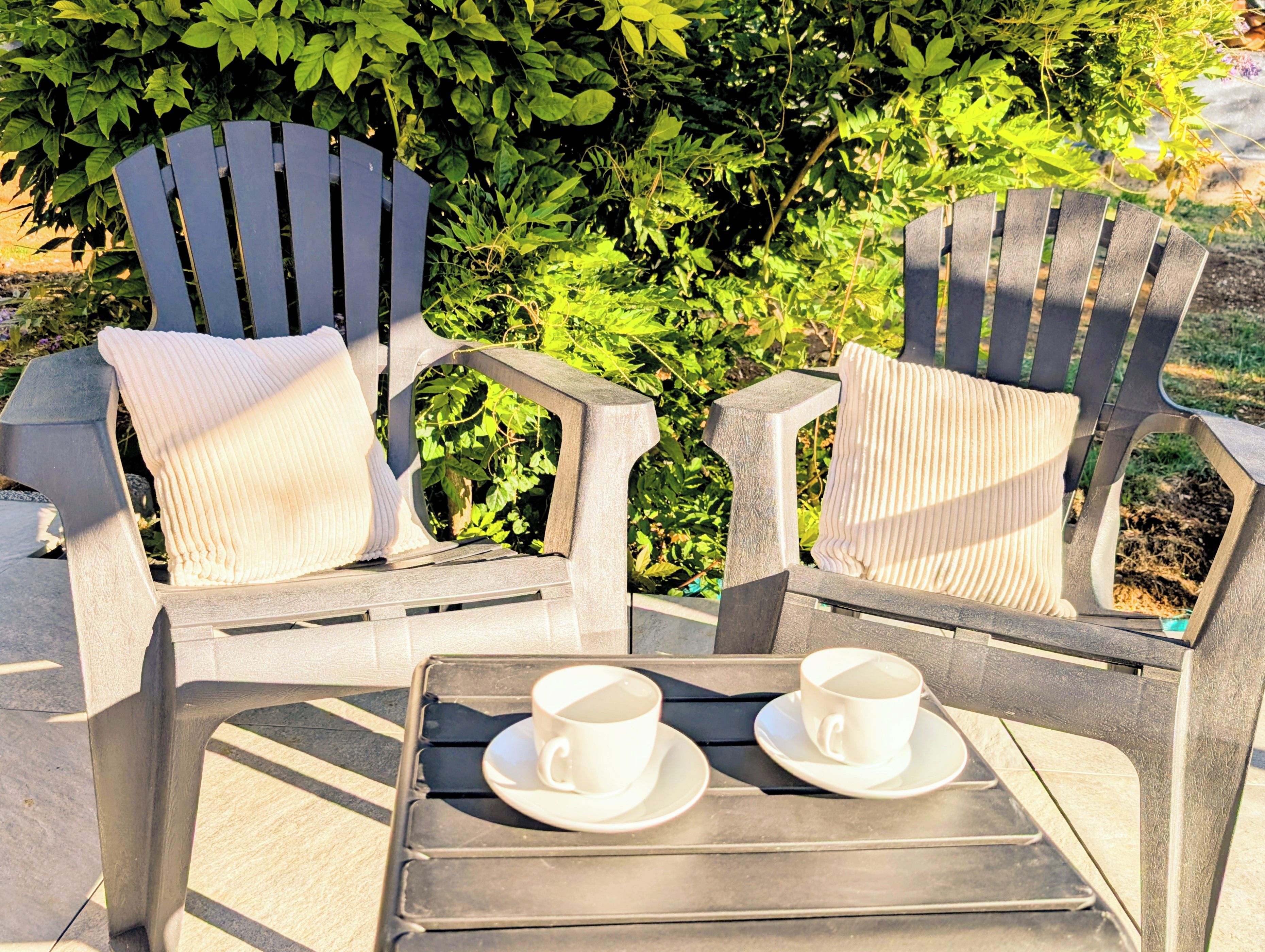 Two Adirondack chairs with white cushions and a tray table with two white cups and saucers.