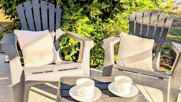 Two Adirondack chairs with white cushions and a tray table with two white cups and saucers.