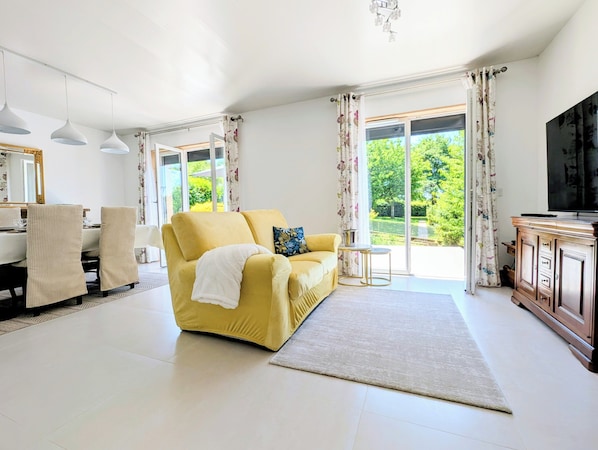 A living room with a yellow sofa, a TV, and a large window with curtains. - 4-Star Comfort Villa with Air Conditioning in a Quiet Location & Beautiful Terrace; Parking 10 Minutes from Besançon (Dannemarie-sur-Crète)