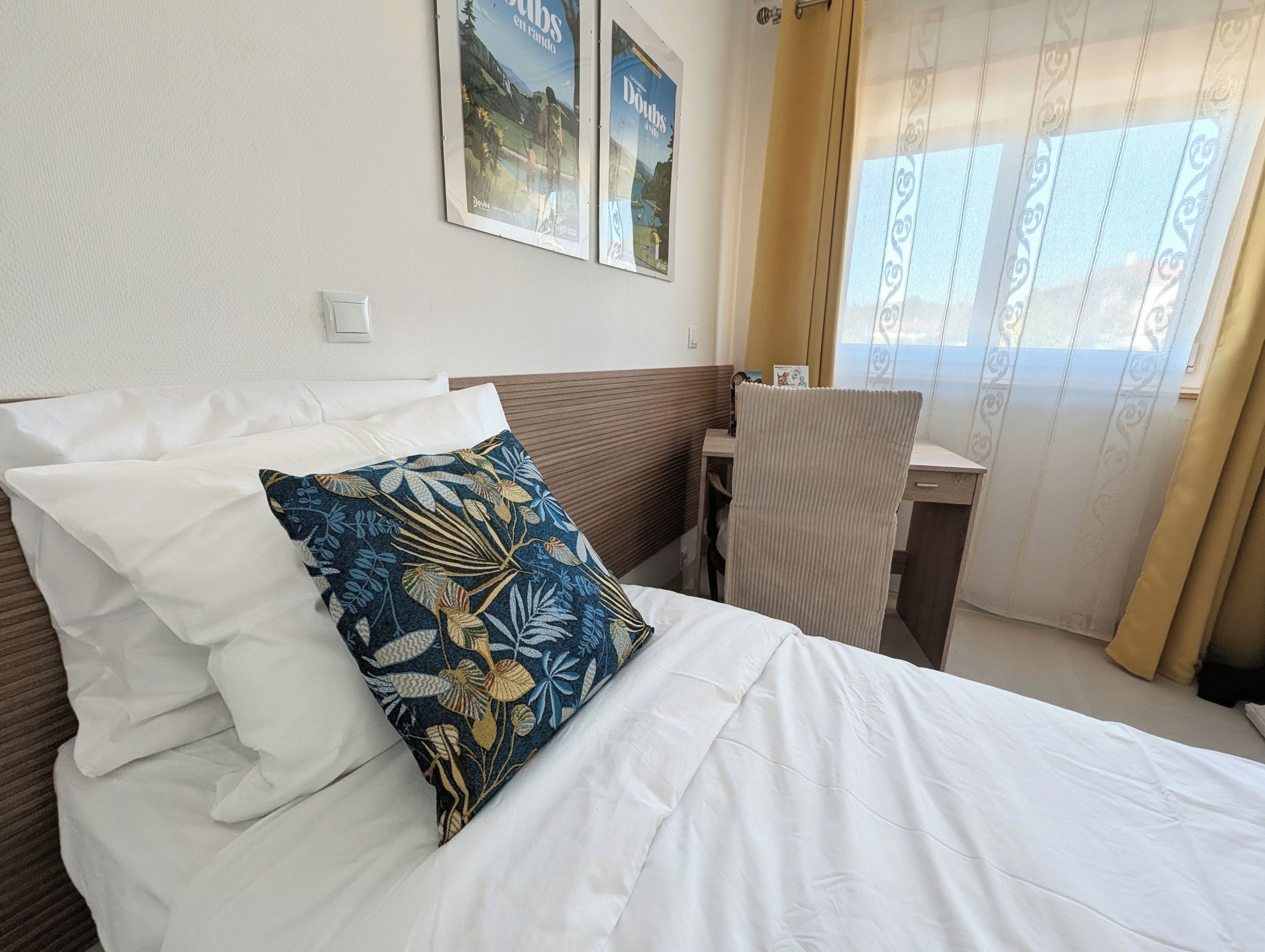 Une chambre d’hôtel avec un lit, un bureau et une chaise. Il y a deux affiches accrochées au mur.