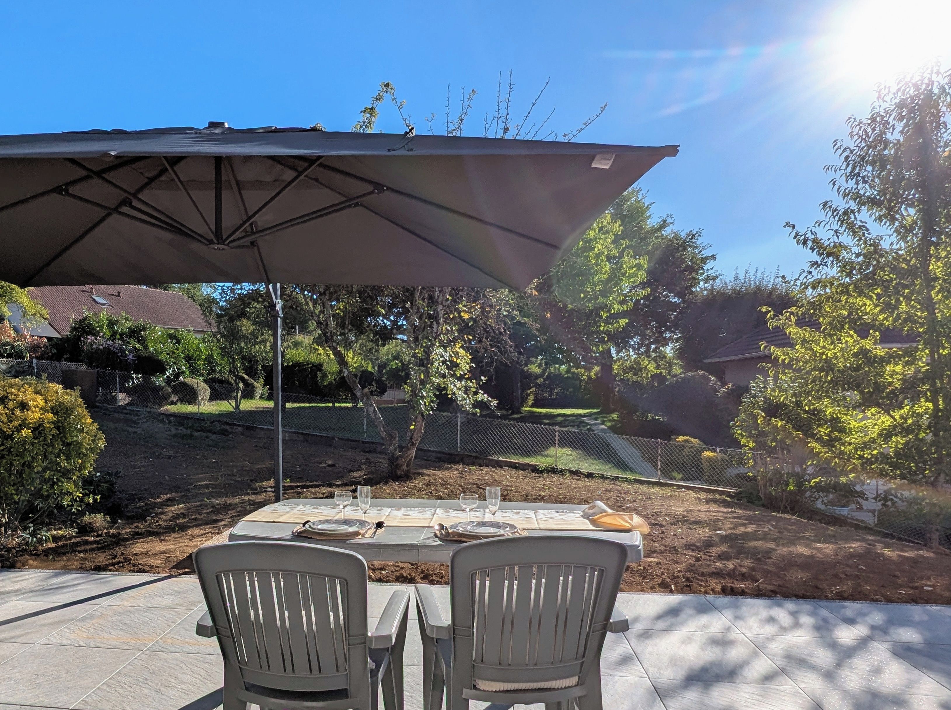 An outdoor patio with a table set for two, a large umbrella, and a view of a backyard with a fence and trees.