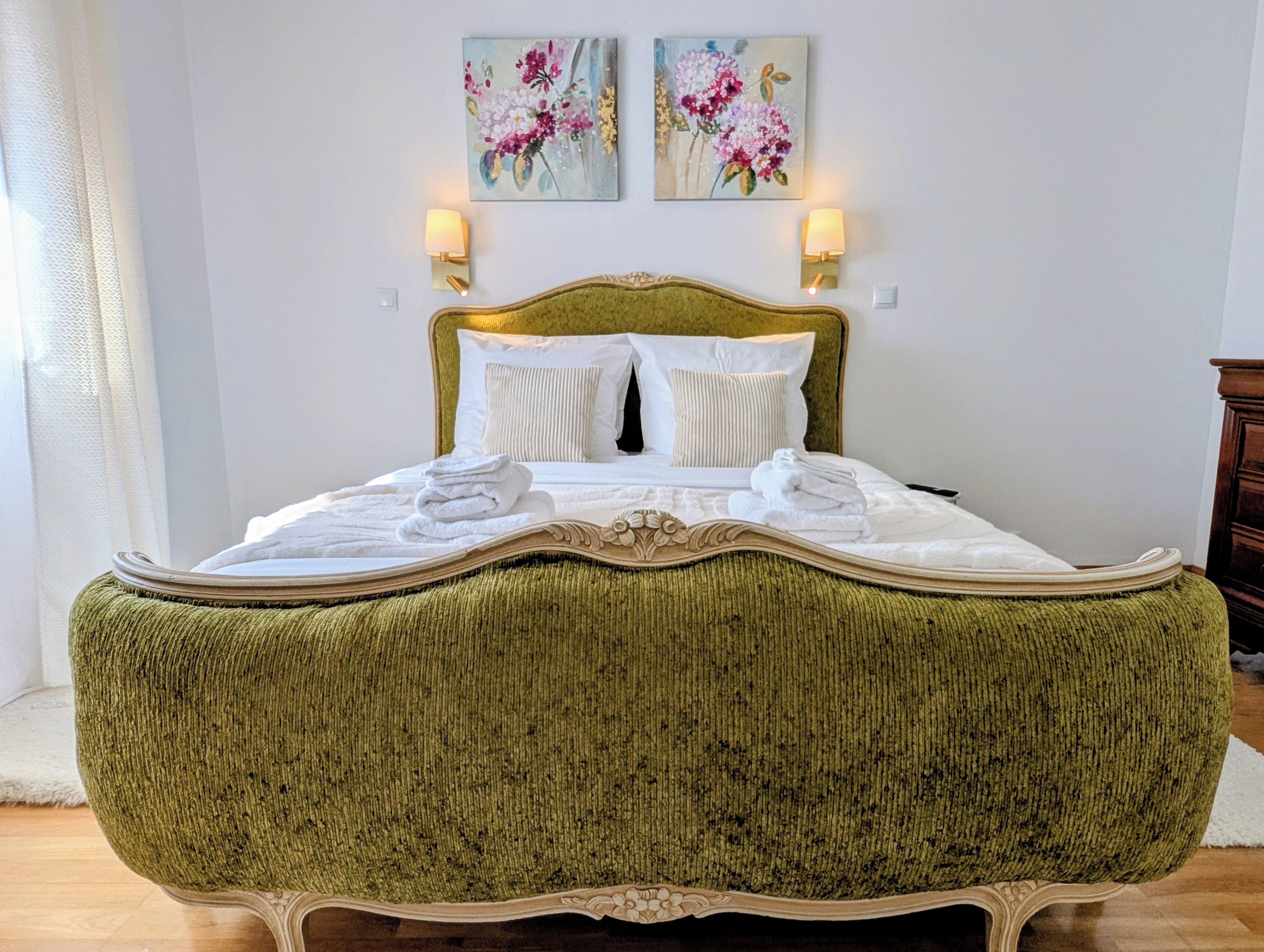 Una cama con cabecera tapizada de verde, ropa de cama blanca y dos lámparas fijadas a la pared.