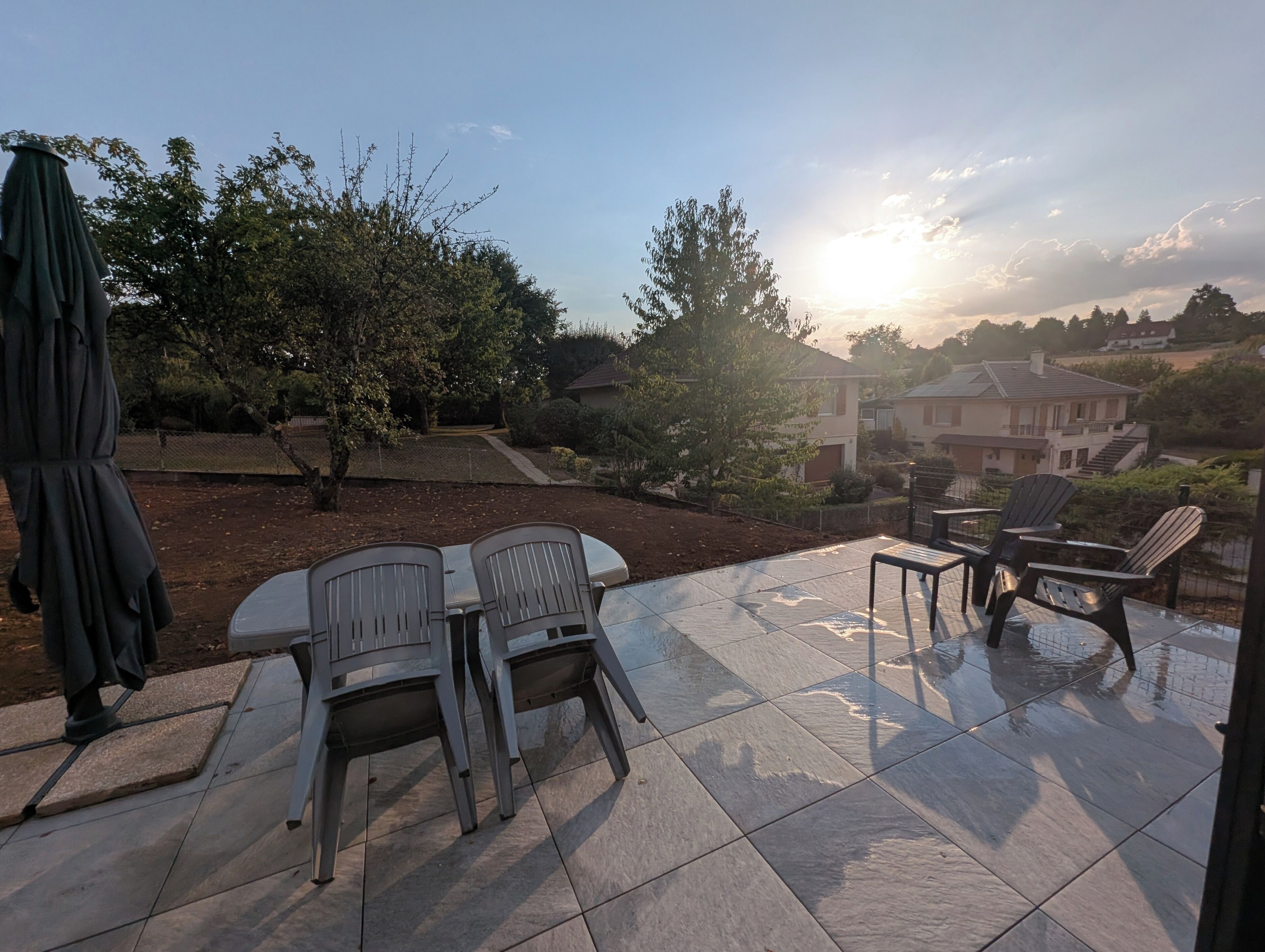 Een terras buiten met een tafel, stoelen, een parasol en huizen op de achtergrond.
