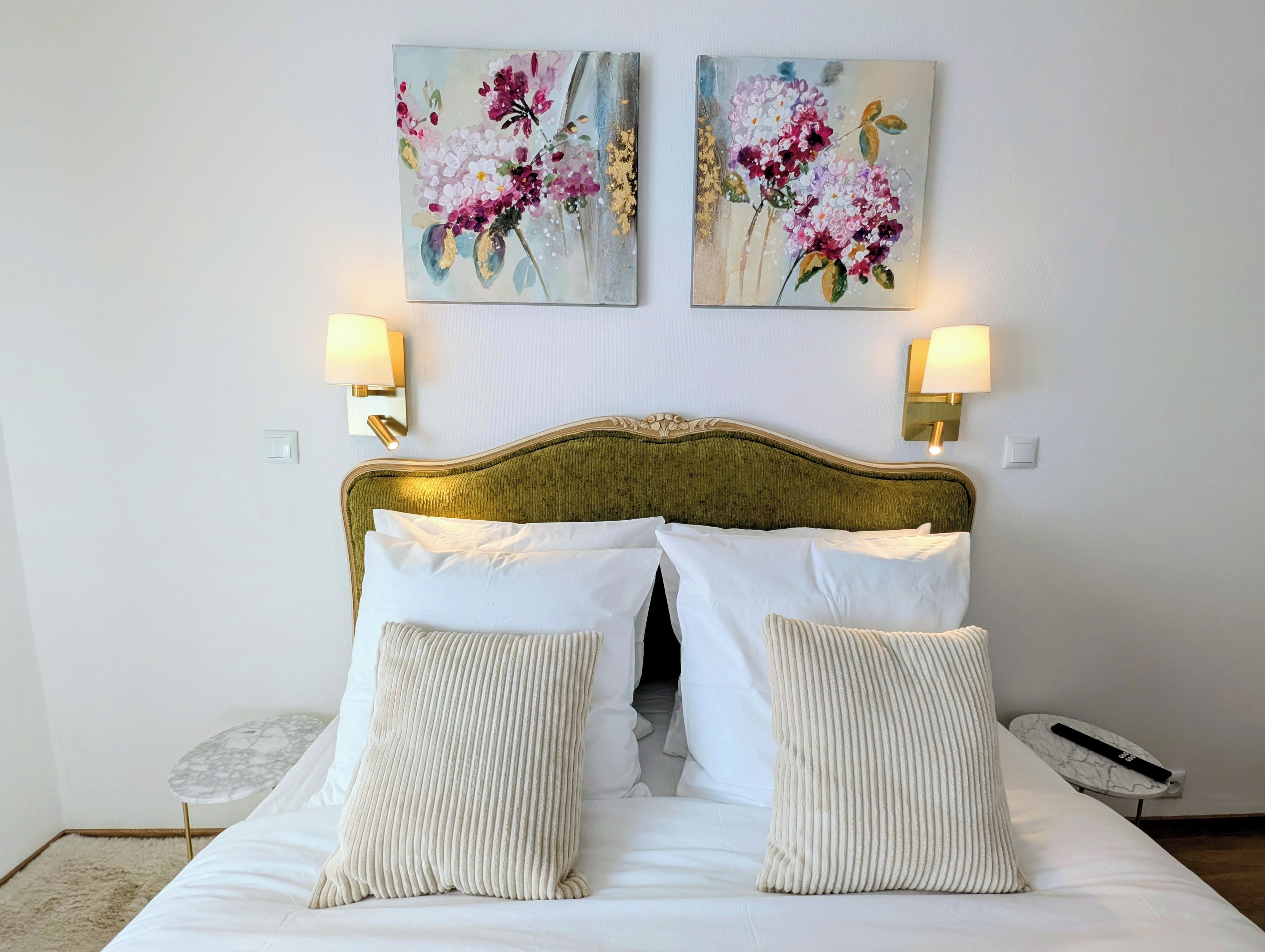 A hotel room with a bed, two wall-mounted lamps, and two floral paintings.