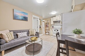 A living room with a sofa, coffee table, and dining table. There is a kitchen area with cabinets and a refrigerator. - Comfy 1 Bedroom Suite by Mroke Vacations (Vancouver)