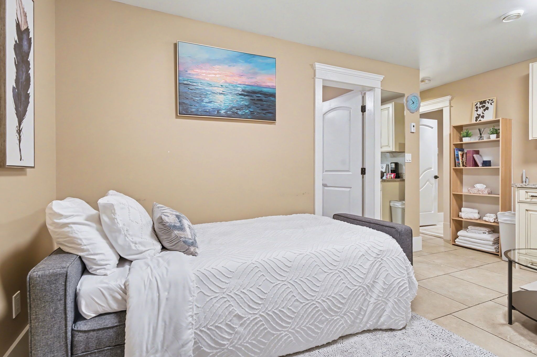 A bedroom with a bed, pillows, a painting, a wardrobe, and a shelf with books and towels.