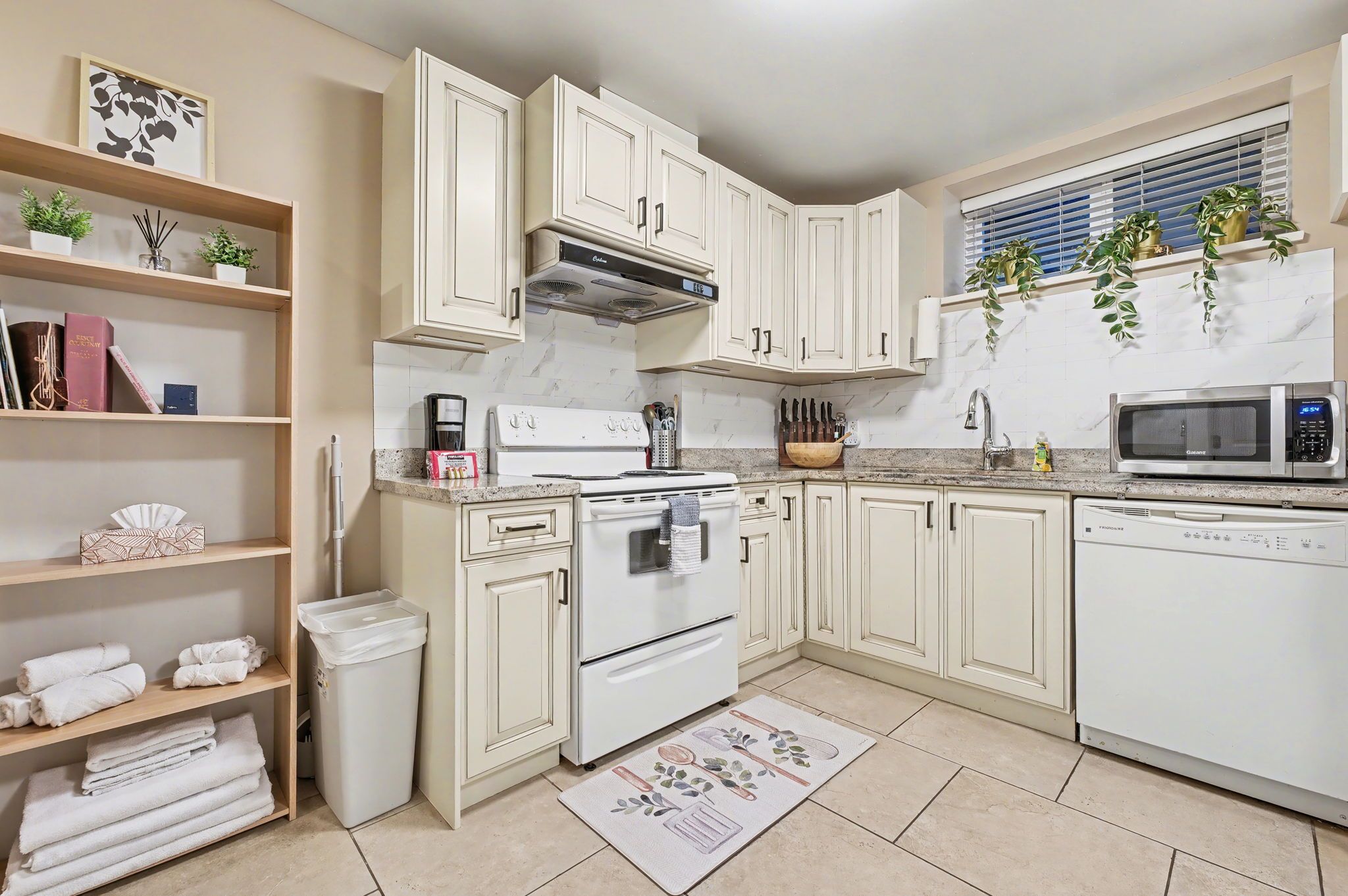 A kitchen with white cabinets, a stove, microwave, and dishwasher. There are shelves with books and plants, and a window with blinds.