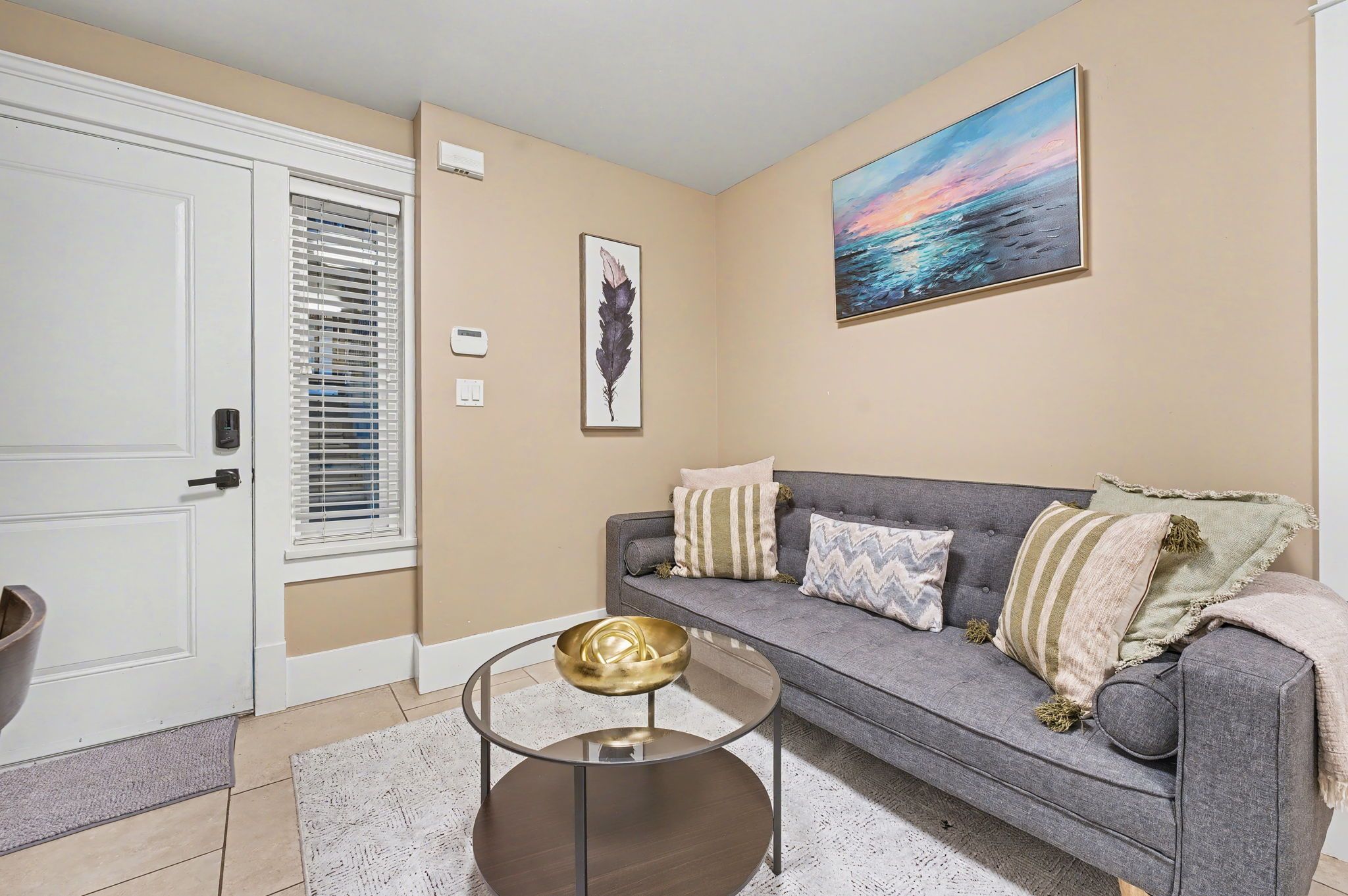A living room with a grey sofa, a glass coffee table, a painting of a sunset, and a door with a window.