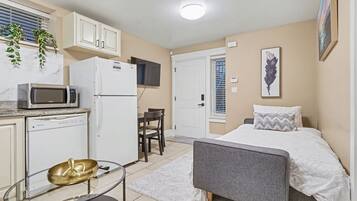 A compact room with a bed, a small kitchenette, a dining area, and a window with blinds.