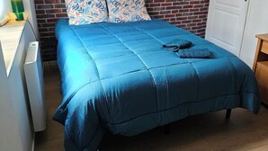 Une chambre avec un mur décoré d’un motif en briques, une couvre-lit bleue, deux coussins à motifs, une lampe de chevet et un plancher en bois.