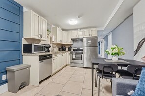 Una cocina moderna con armarios blancos, un refrigerador de acero inoxidable, un microondas y un lavavajillas. Hay una mesa de comedor con sillas y una planta en maceta.