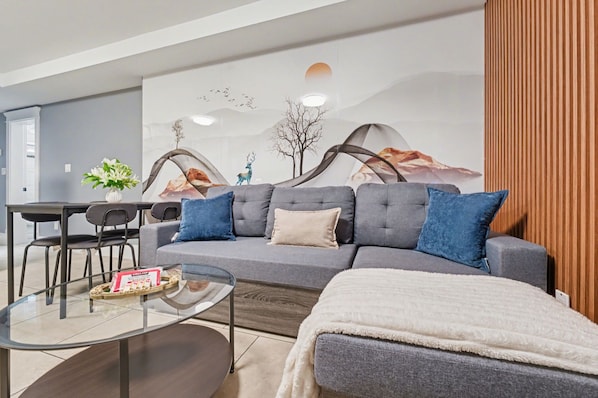 A modern living room with a grey sofa, blue cushions, a glass coffee table, and a white throw blanket.