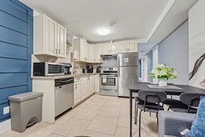 A modern kitchen with white cabinets, a stainless steel refrigerator, a microwave, and a dishwasher. There is a dining table with chairs and a potted plant. - Quiant 2 Bedroom Suite (Vancouver)