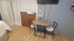 Una habitación pequeña con una cama, una mesa con sillas, un televisor montado en la pared y un ventanal con persianas.
