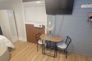 A small room with a bed, a table with chairs, a TV mounted on the wall, and a window with blinds. - Secluded Bachelor Suite by Mroke Vacations (Vancouver)