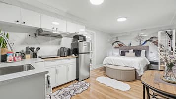 A modern kitchen with white cabinets, a stainless steel refrigerator, and a microwave. There is a dining area with a wooden table and chairs, and a bedroom with a bed and a nightstand.