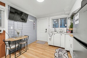 A compact kitchen with a dining table, a TV mounted above it, and a refrigerator. - Secluded Bachelor Suite (Vancouver)
