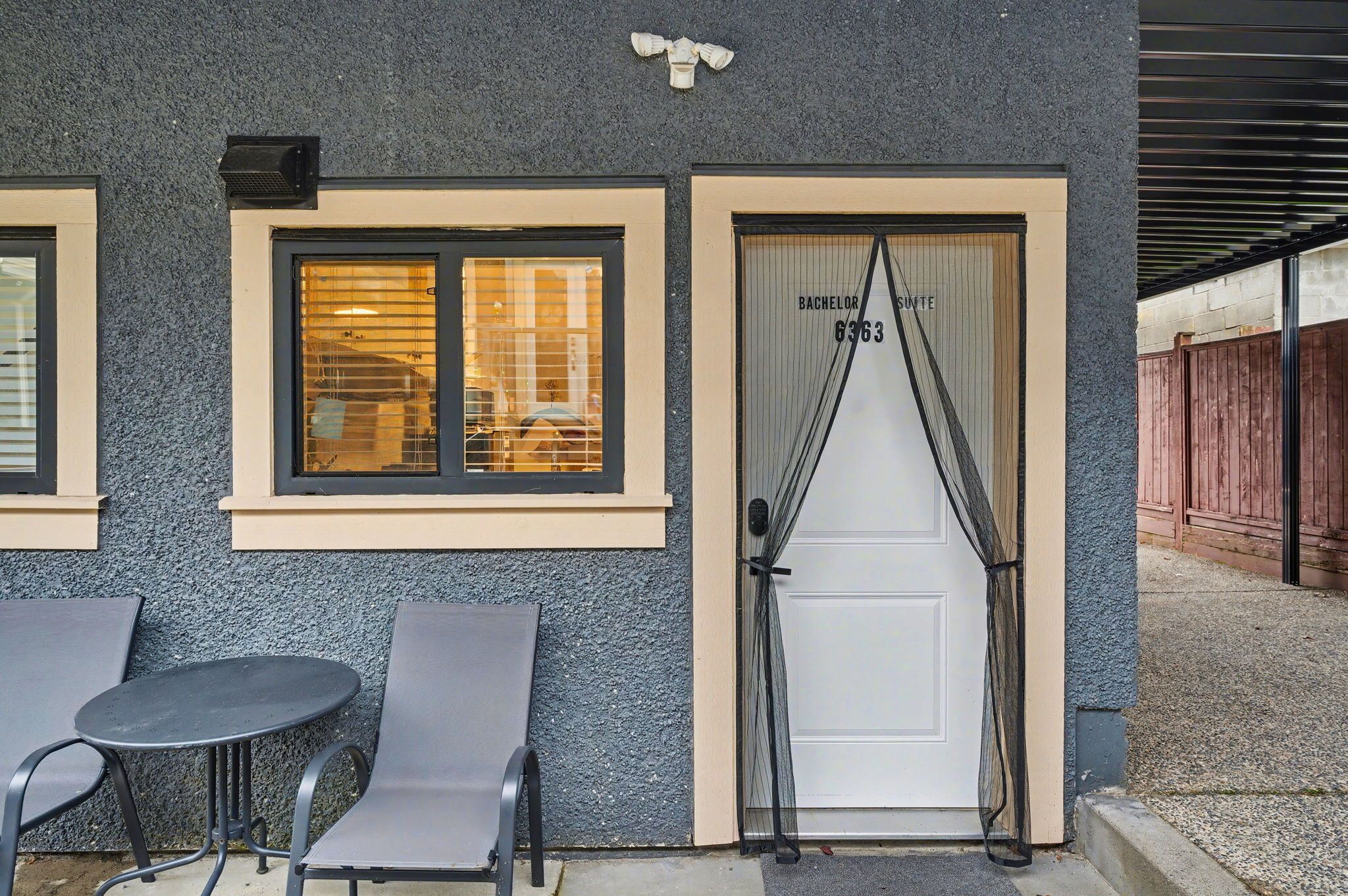 A building exterior with a textured facade, a door labeled 'Bachelor Suite 6363,' and two outdoor tables with chairs.