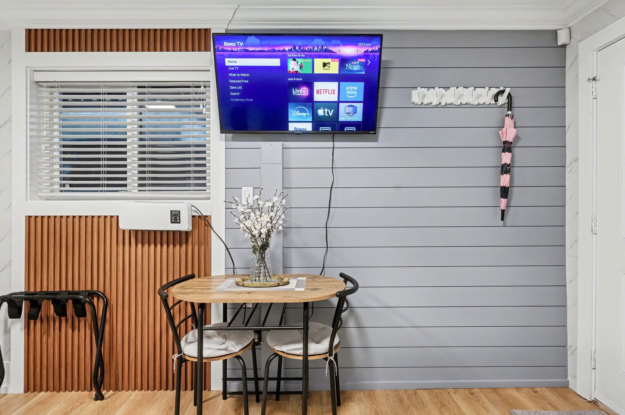 A room with a TV mounted on the wall displaying a streaming service interface, a wooden table with chairs, a window with blinds, and a wall-mounted rack with an umbrella.