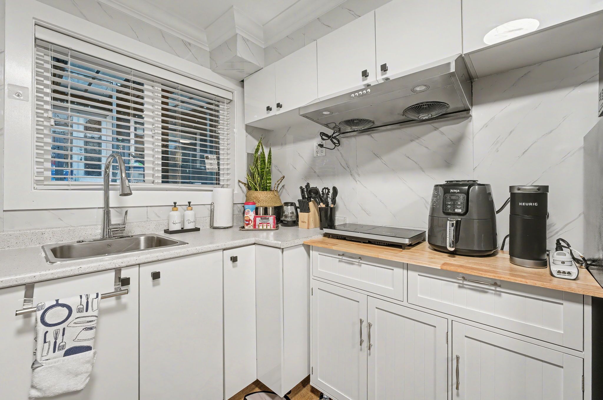 A modern kitchen with white cabinets, a stainless steel sink, a window with blinds, a coffee maker, and a kettle.