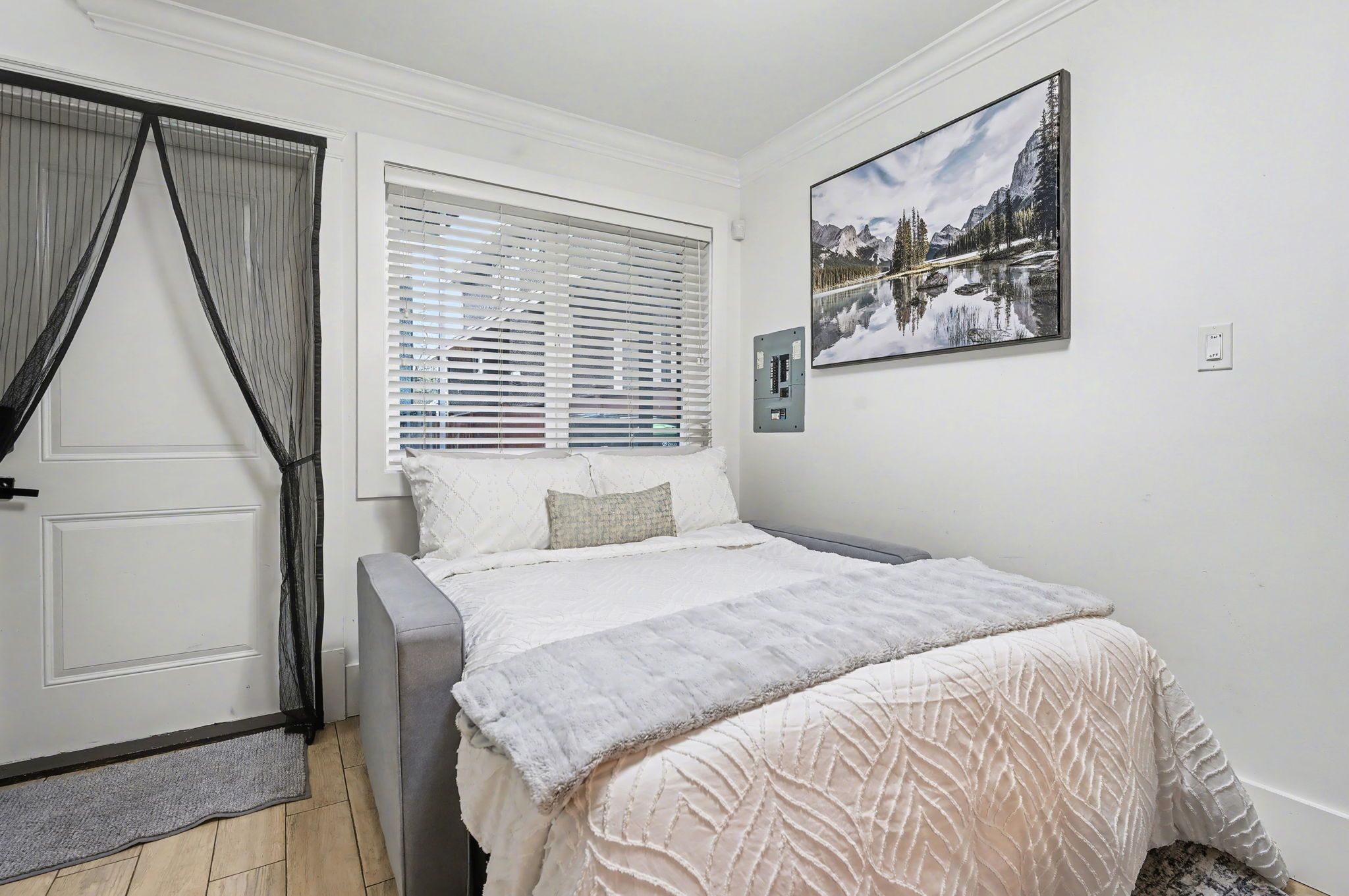 Une chambre à coucher avec un lit, une fenêtre avec des stores, une porte munie d’une moustiquaire et un tableau accroché au mur.