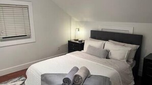 Ein ordentlich gemachtes Bett mit Kissen und Decke, ein Nachttisch mit Lampe und ein Fenster mit Jalousien.