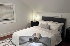 Una cama bien hecha con almohadas y una colcha, una mesita de noche con una lámpara y una ventana con persianas.