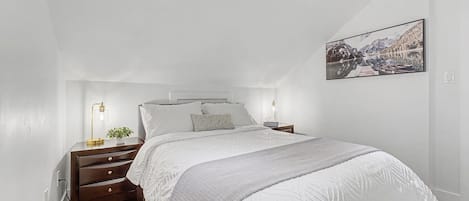 A bedroom with a bed, bedside tables, a nightstand, a wall-mounted picture, and a wooden floor.