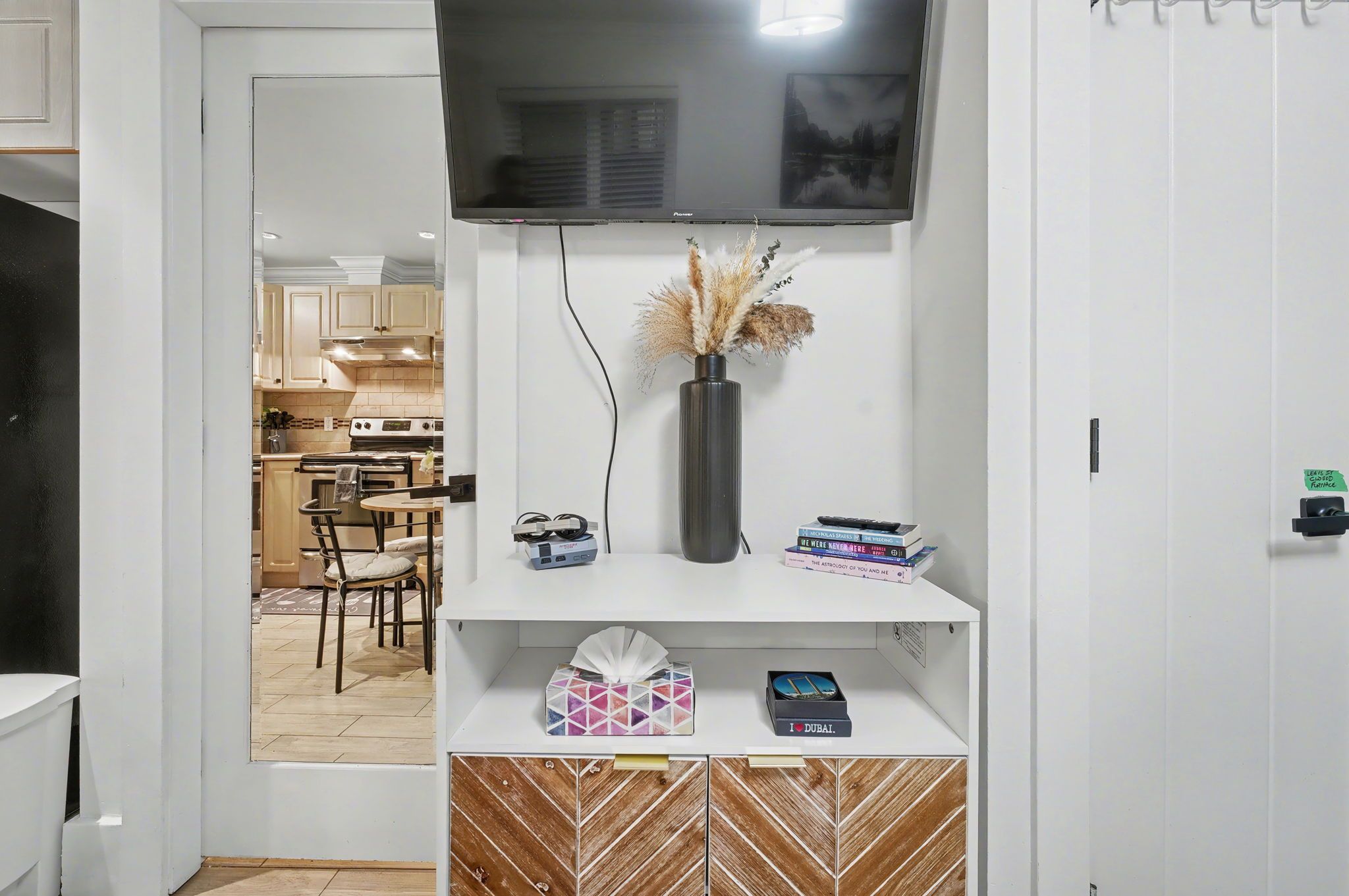 A modern kitchen with a flat-screen TV, a vase with dried plants, and a dining area with chairs and a table.