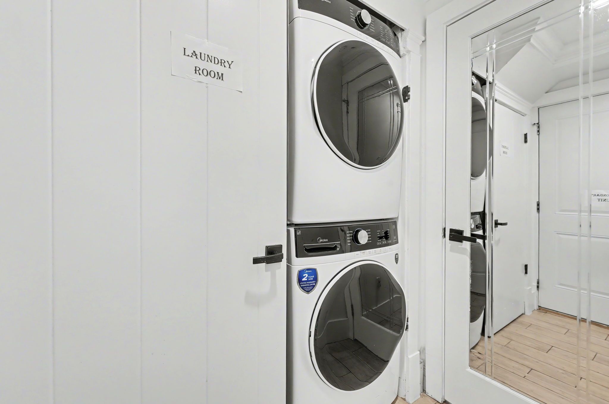 A laundry room with two stacked washing machines, a sign indicating the room, and a door leading to another area.