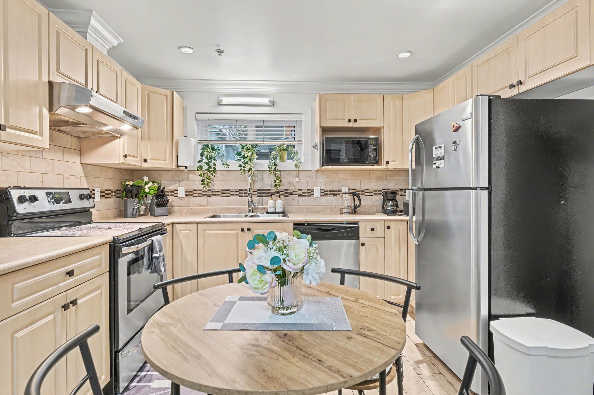 A modern kitchen with wooden cabinets, stainless steel appliances, a dining table with chairs, and a vase with flowers.