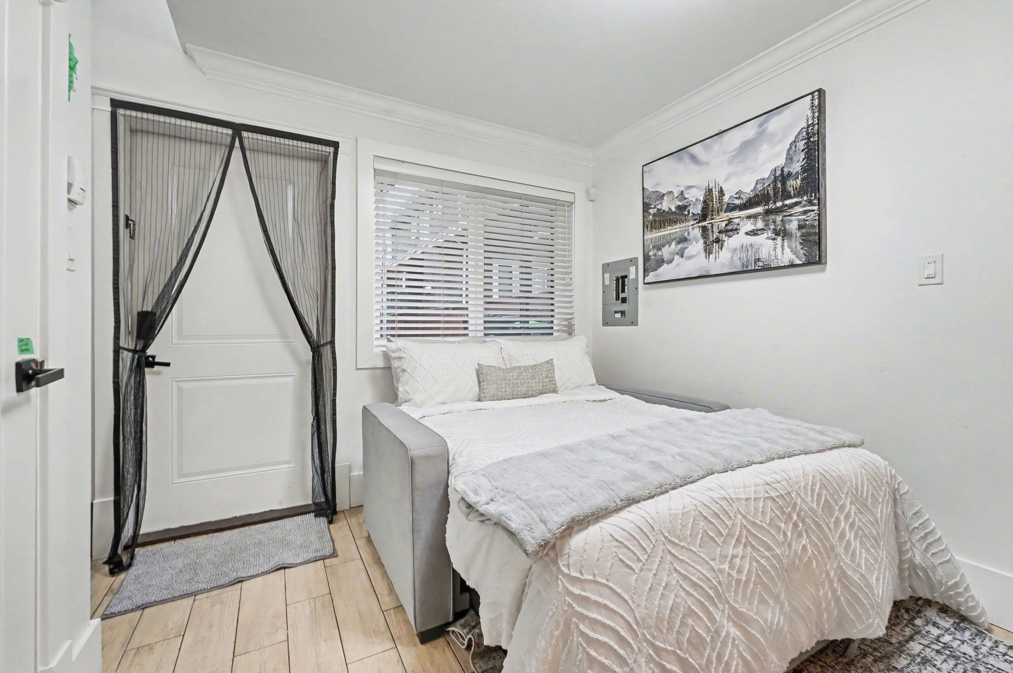Ein Schlafzimmer mit Bett, einer Tür mit Vorhängen, einem Fenster mit Jalousien und einem Wandbild.
