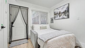 Ein Schlafzimmer mit Bett, einer Tür mit Vorhängen, einem Fenster mit Jalousien und einem Wandbild.