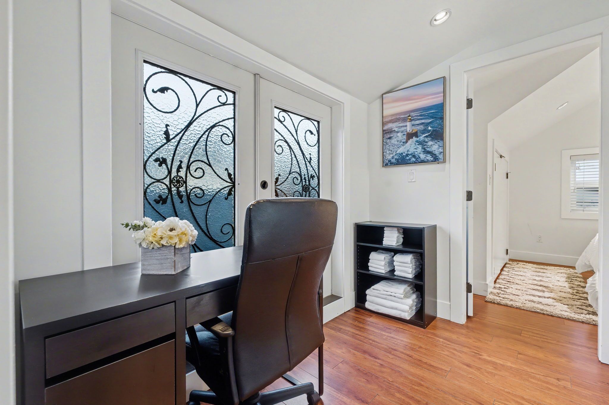 A room with a desk, chair, decorative window, shelves with folded towels, and a painting on the wall.