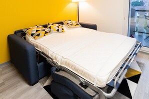 Ein schmales Bett mit Matratze und Kissen, ein Nachttisch, eine Lampe und ein Blick durch das Fenster nach draußen.