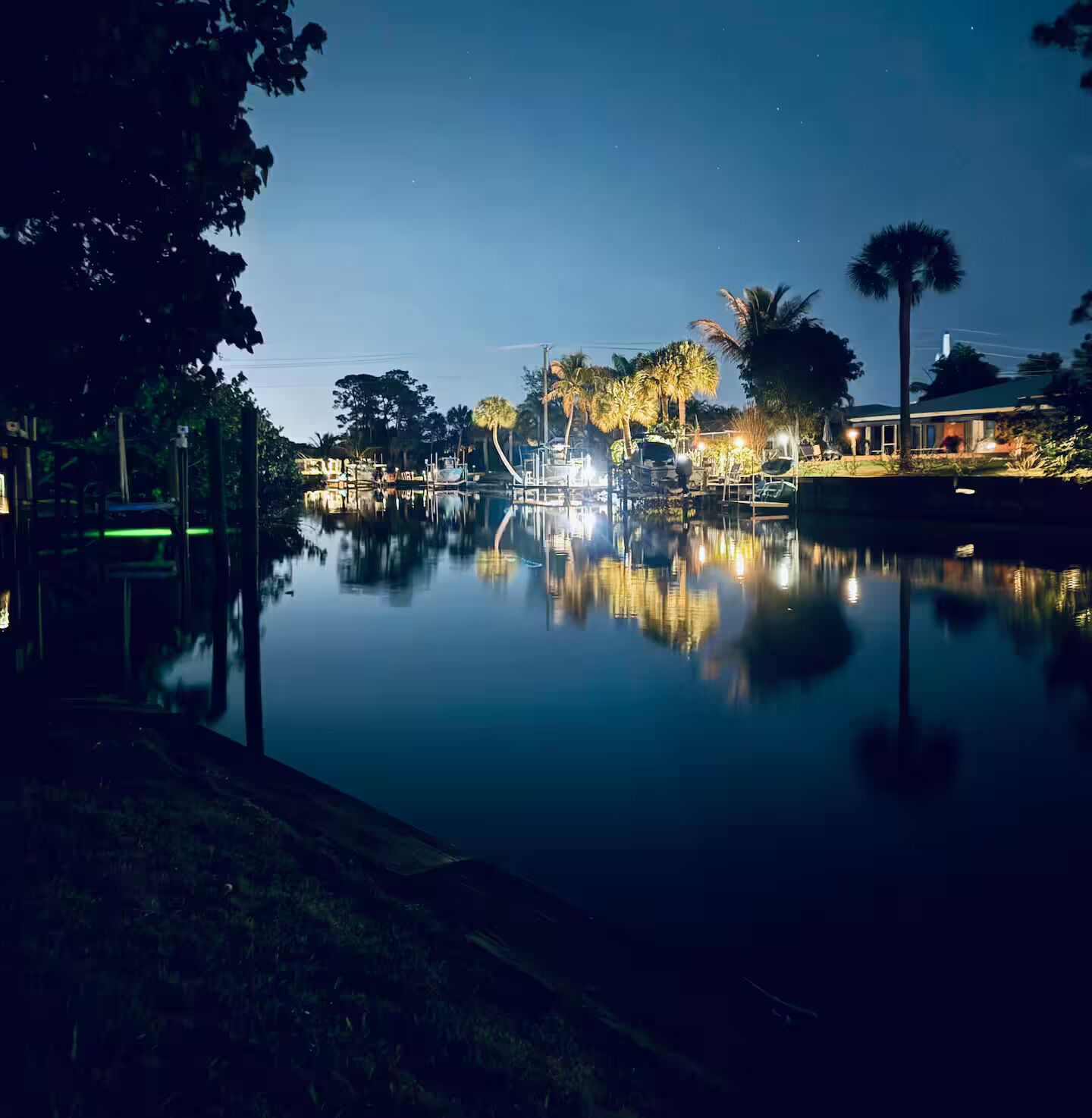 Vista nocturna de una masa de agua tranquila que refleja luces, rodeada de árboles y un edificio con un balcón visible.