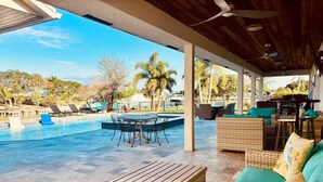 A covered outdoor area with a pool, wicker furniture, and a view of palm trees and a distant building. - Bring Boat! Heated Pool & Spa/Kayaks/Bikes/Fishing (Stuart)