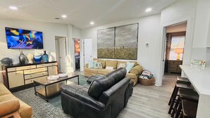 Ett modernt vardagsrum med en svart lädersoffa, en beige soffa, ett soffbord och en tv som är monterad på väggen. Det finns också en matplats med ett vitt bord och stolar.