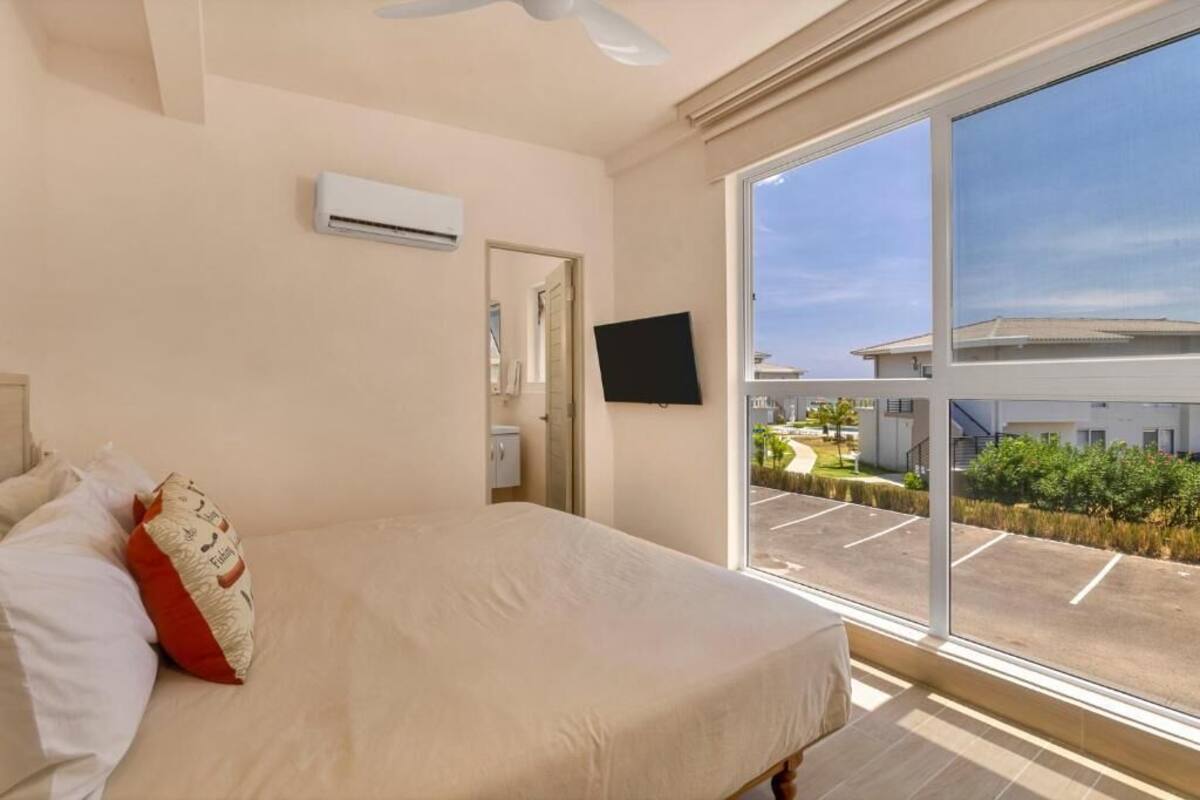 A bedroom with a bed, an air conditioning unit, a TV, and a large window with a view of a parking lot and buildings.