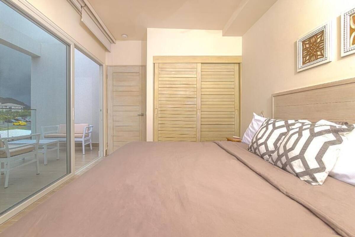 A bedroom with a bed, a sliding glass door leading to a balcony, a wooden wardrobe, and a wall with framed artwork.