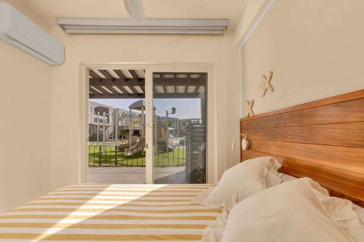 A bedroom with a wooden headboard, a striped bedspread, and a view of a playground through a glass door.