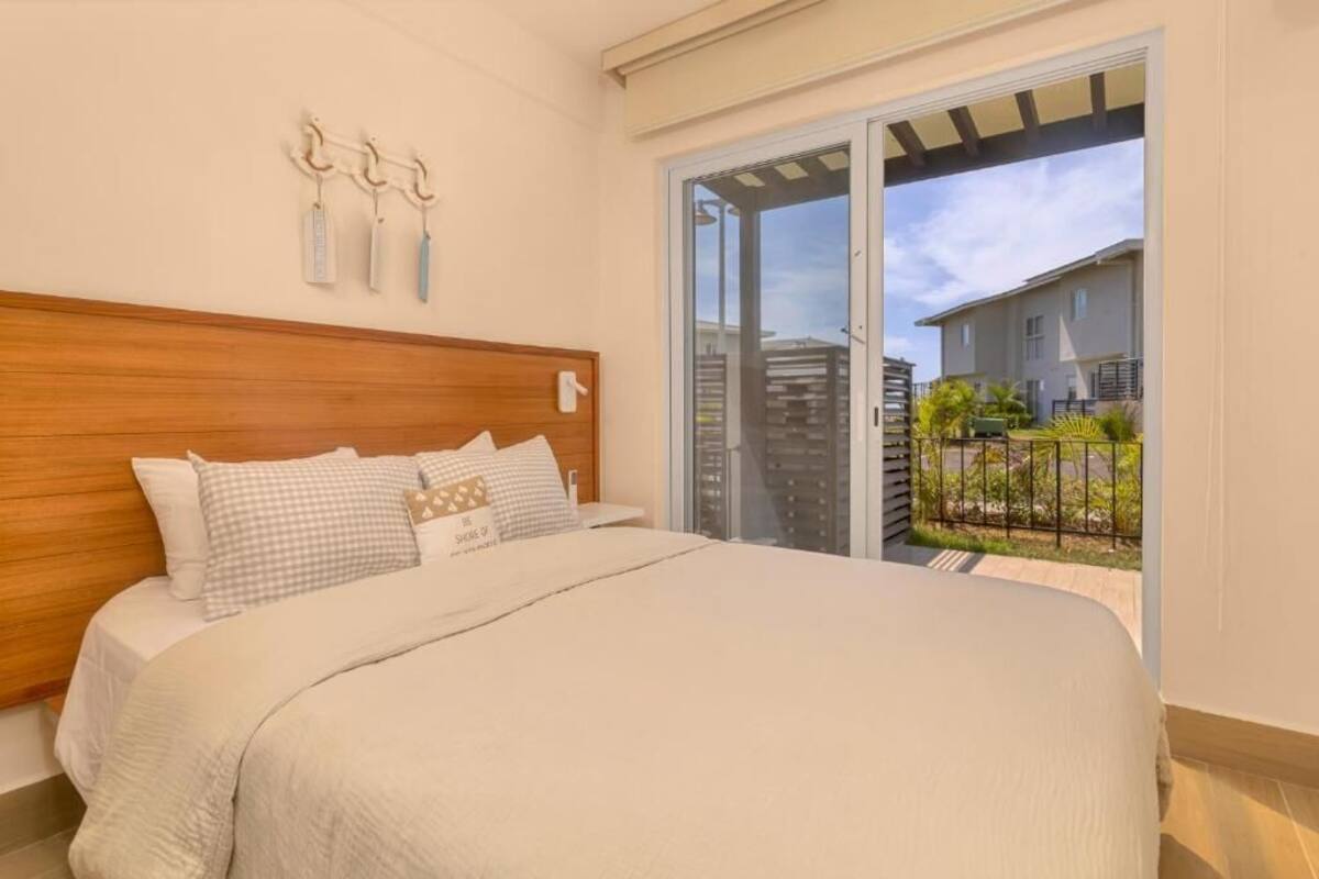 A bedroom with a wooden headboard, a bed with white bedding, and a view of a balcony with a railing and greenery.