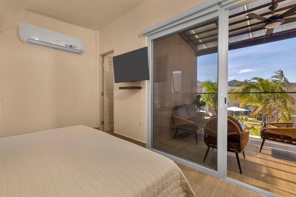Un dormitorio con cama, televisor montado en la pared y puerta corrediza de vidrio que da a un espacio exterior con mobiliario de mimbre y vistas a palmeras.