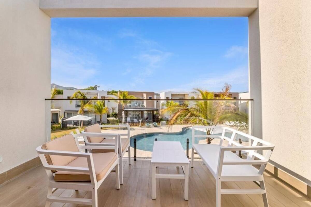 A balcony with a pool view, white furniture, and a clear sky.