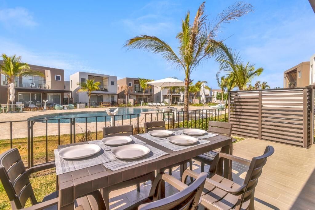Area pranzo a bordo piscina con tavolo apparecchiato per quattro, circondato da sedie, con piscina e palme sullo sfondo.