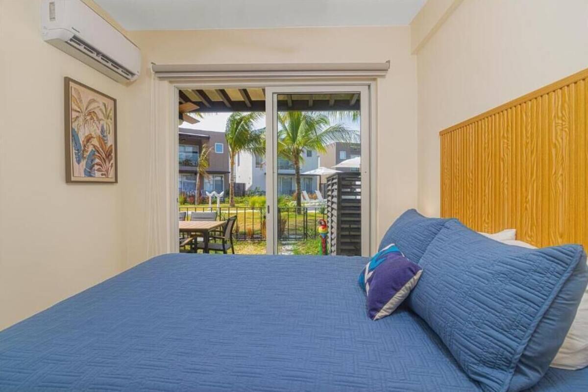 A bedroom with a large bed, a wooden headboard, a sliding glass door leading to a patio with outdoor furniture and palm trees.