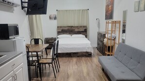 Een kleine kamer met een bed, een slaapbank, een eettafel met stoelen, een aan de muur bevestigde televisie en een kitchenette.