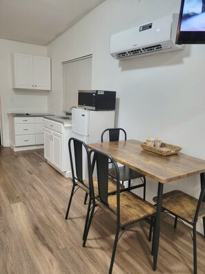 A compact kitchen with white cabinets, a microwave, and an air conditioner. There is a dining table with chairs and a basket of items on it.