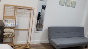Een kamer met een grijze bank, een houten plank met opgevouwen dekens, een spiegel en drie ingelijste foto's aan de muur.